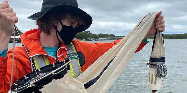 Hydrotech standing in a boat, wearing safety orange jacket and flotation vest, lowering fine mesh net over the side of the boat