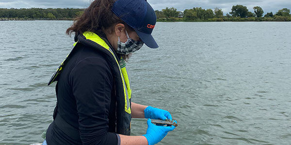 Scientist standing in a boat (only the lake and shoreline are visible), wearing blue latex gloves and holding a passive ZM sampler