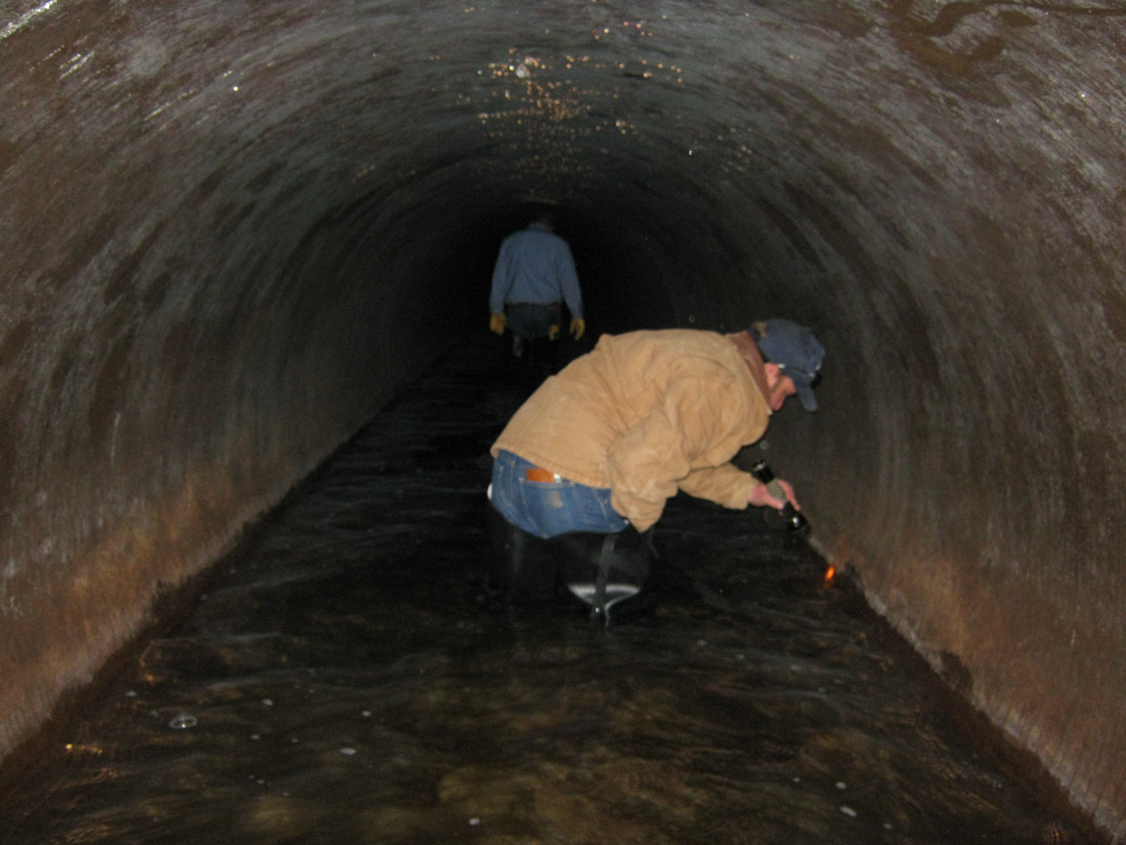 Scientist crouching in a drained, large water supply pipe looking for juvenile or adult zebra mussels along the pipe walls.   Another scientist is visible in the background walking further down the pipe.