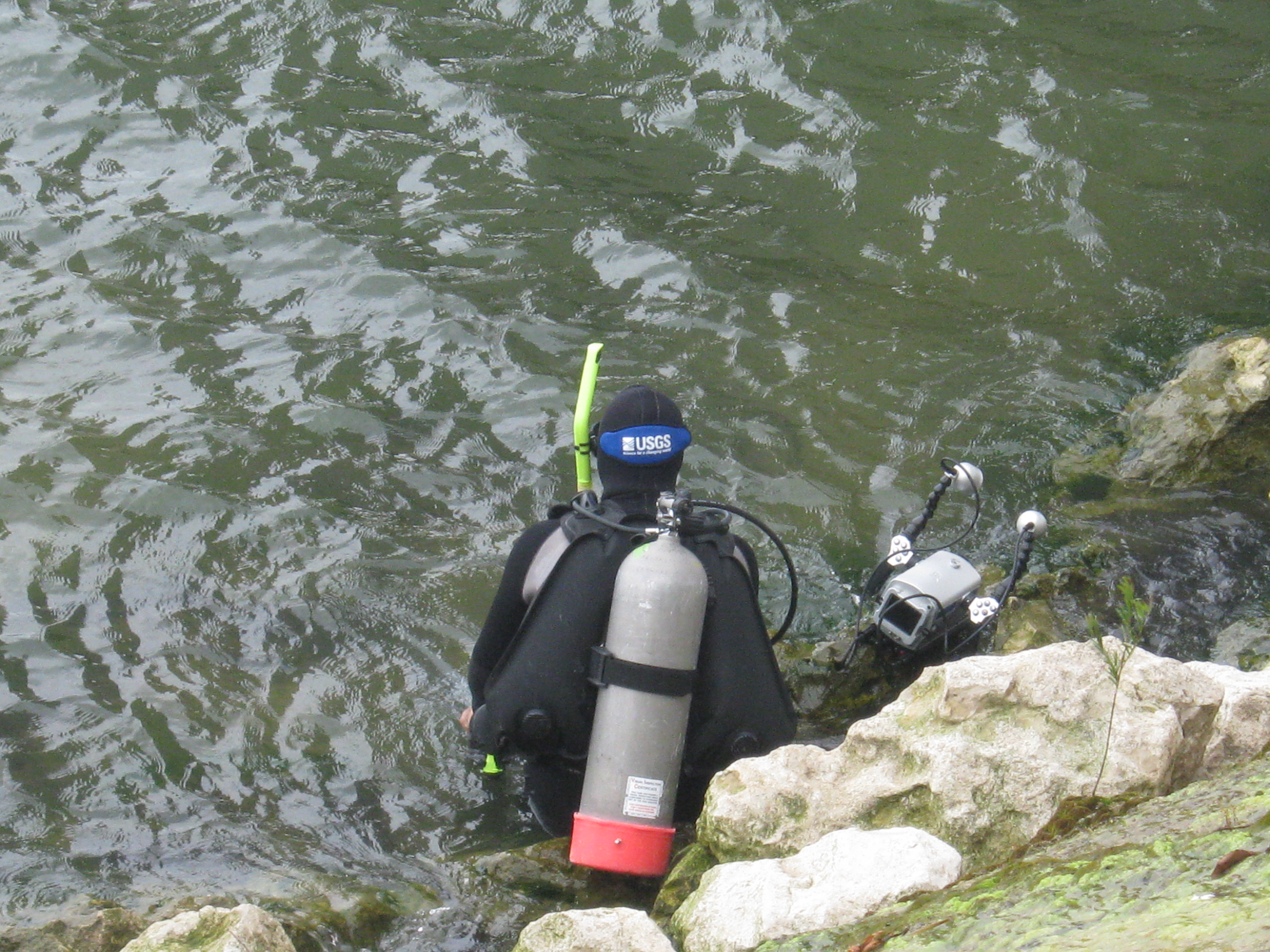 Scuba diver at a rocky shoreline entering the water, holding an underwater camera
