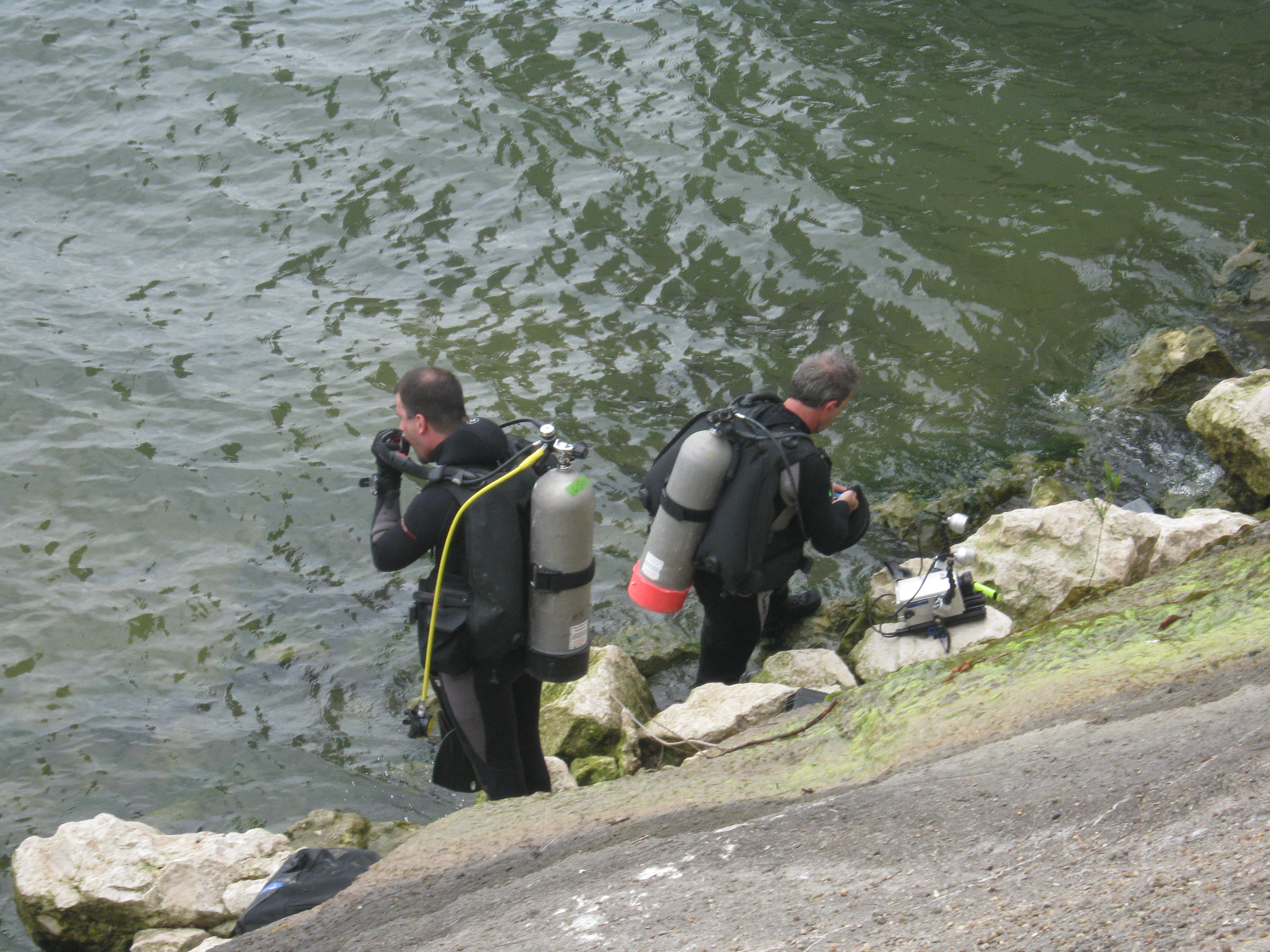 Two scuba divers, standing in the water along the rocky lake edge, putting on their dive masks
