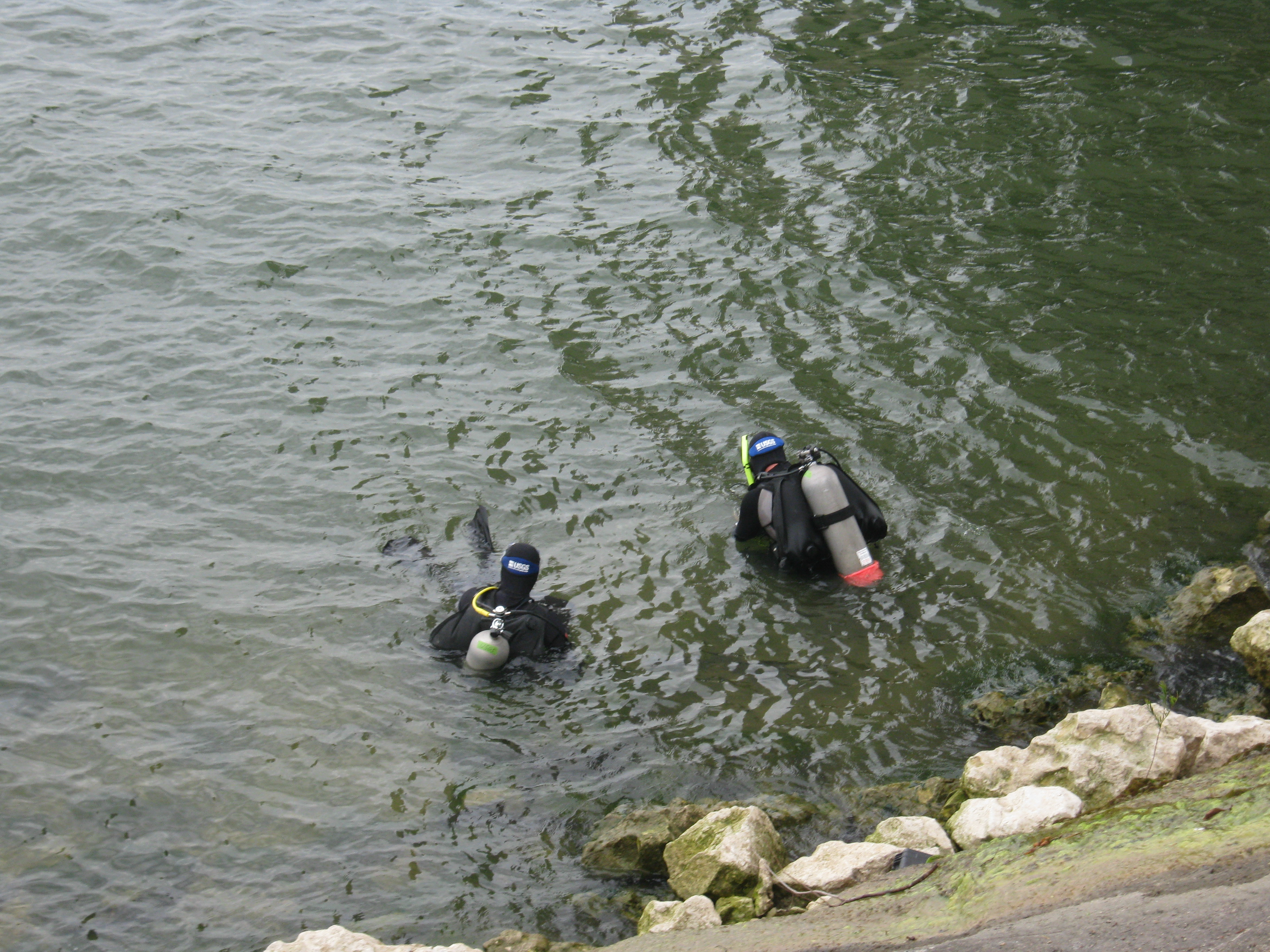 Two scuba divers, in the water a few feet from the rocky lake edge, beginning to swim