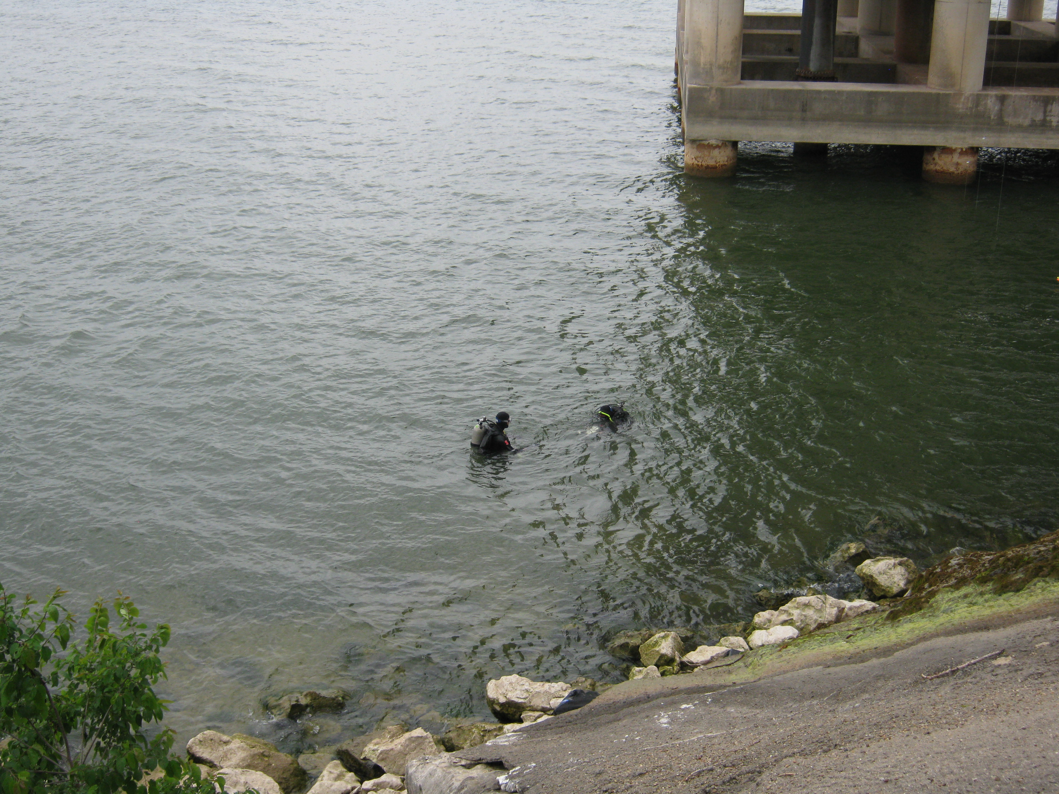 Two scuba divers, swimming across the surface of the water towards a large concrete structure in the lake.