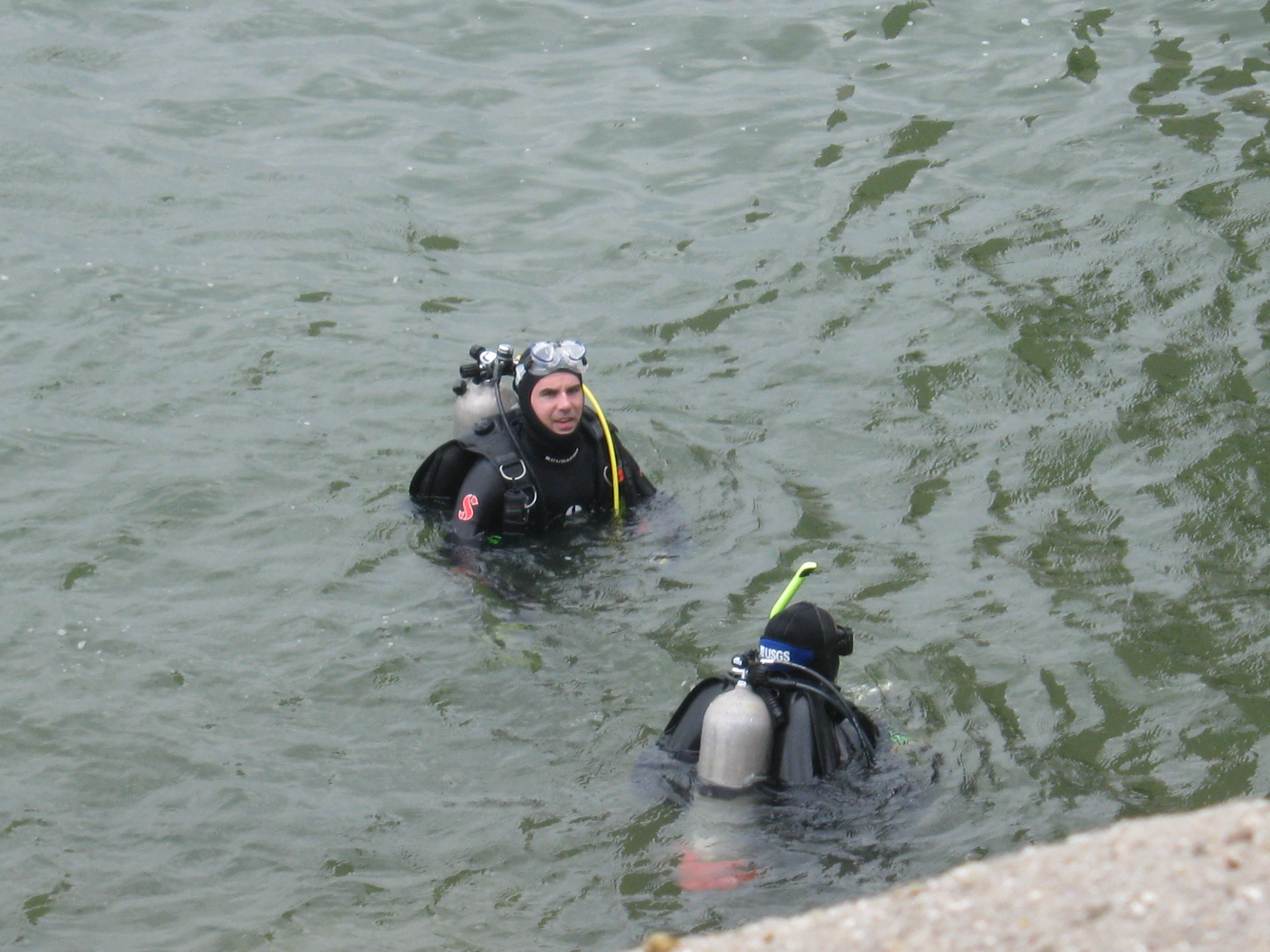 Two scuba divers, head and shoulders above the water, with the lake shore visible in the bottom corner of the image
