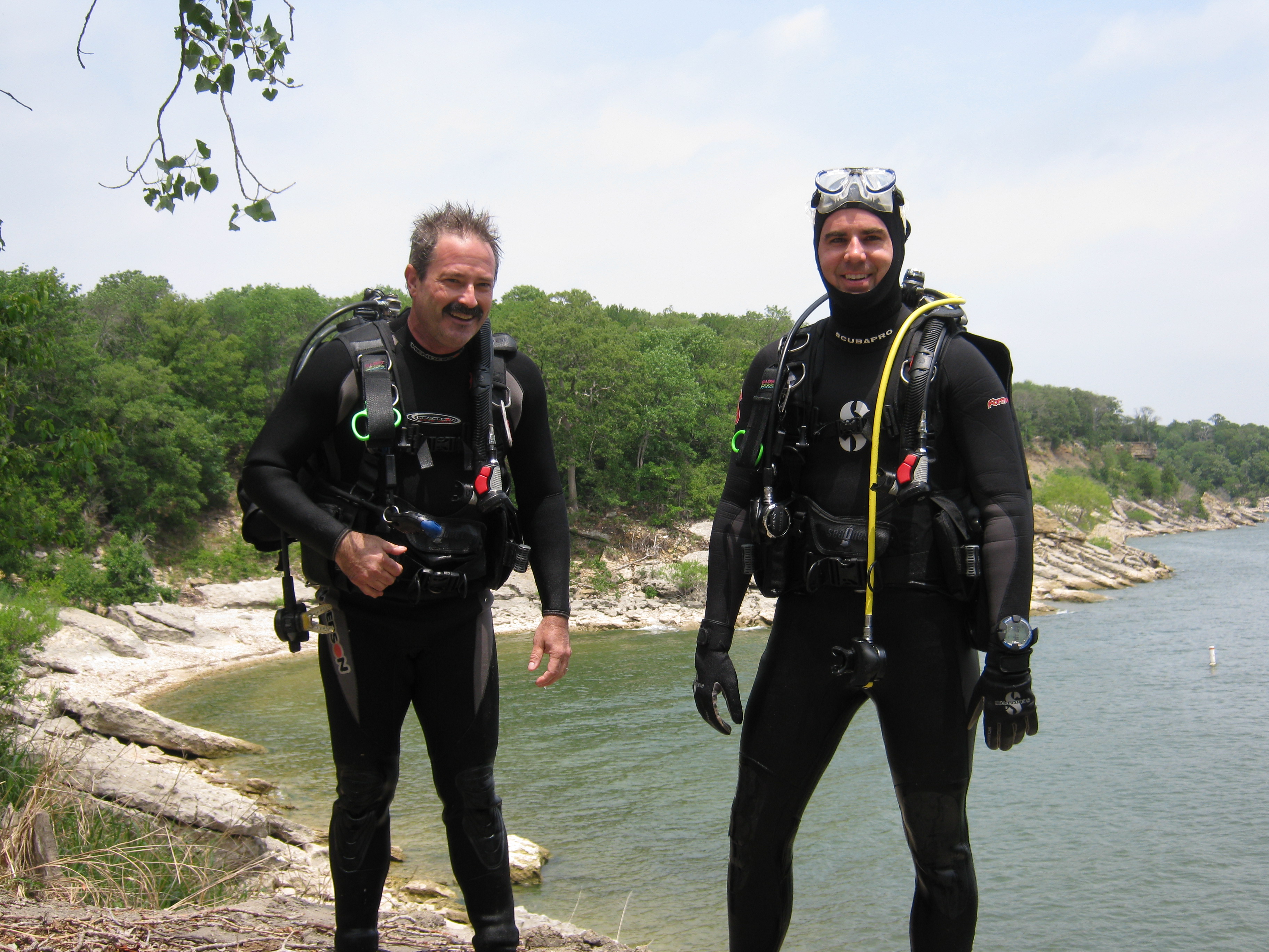 Two divers in full scuba gear, standing and facing the camera.  Lake and shoreline behind them.